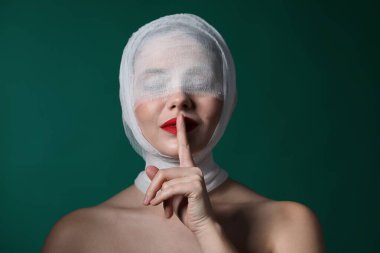 Estetik ameliyattan sonra yüzü bandaja sarılı bir kadın yeşil arka planda sessizlik jesti gösteriyor.