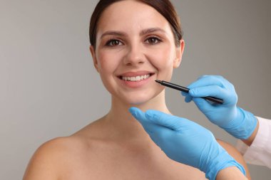 Doktor, gri arkaplana karşı estetik ameliyat için hastanın yüzüne işaretler çiziyor.