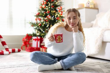 Süveter giymiş, elinde bir fincan sıcak içecekle gülümseyen bir kadın Noel için dekore edilmiş odanın bulanık ışıklarına karşı. Metin için boşluk