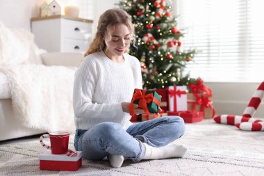 Evde bulanık ışıklara karşı elinde Noel hediyeleriyle gülümseyen bir kadın.