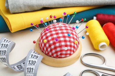Checkered pincushion with pins, spools of threads, fabrics and measuring tape on light table, closeup