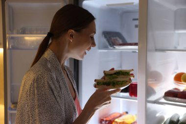 Mutfakta açık buzdolabının yanında sandviçleri olan bir kadın.