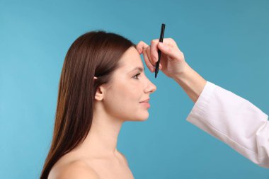 Doktor, açık mavi arkaplana karşı estetik ameliyat için hastanın yüzüne işaretler çiziyor.