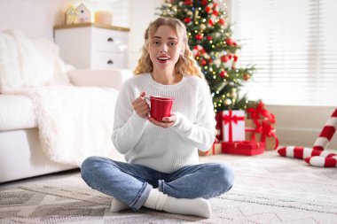 Süveter giyen, elinde bir bardak sıcak içecekle gülümseyen kadın Noel için dekore edilmiş odadaki bulanık ışıklara karşı.