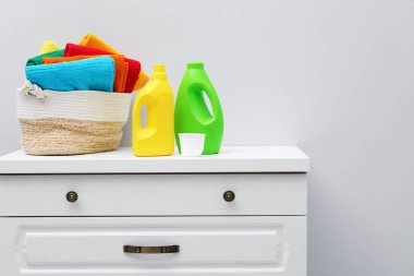 Çamaşır sepetinin içinde havlular ve deterjan şişeleri beyaz duvarın yanındaki dolapta.