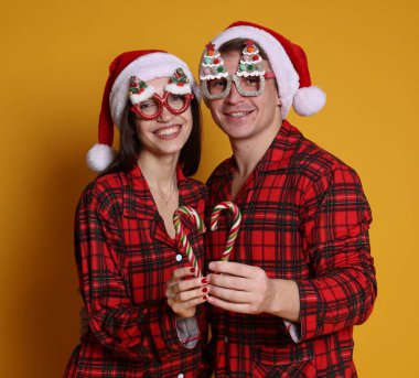 Noel Baba şapkalı ve pijamalı mutlu çift turuncu arka planda şeker kamışlarıyla kalp yapıyorlar. Noel atmosferi