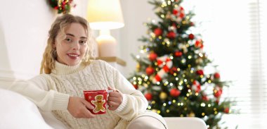 Süveter giymiş, elinde bir fincan sıcak içecekle gülümseyen kadın evdeki bulanık ışıklara karşı koltukta dinleniyor, mesaj için yer var. Mutlu Noeller ve mutlu yıllar.