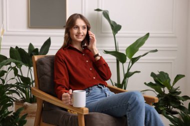 Elinde bir fincan aromatik kahve olan kadın evdeki koltukta telefonla konuşuyor. Metin için boşluk