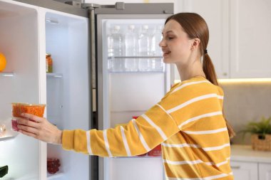 Kadın mutfaktaki buzdolabının rafına yiyecek koyuyor.
