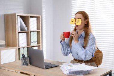 Yapışkan notlara sahte gözleri çizilmiş bir kadın bir bardak içki tutuyor ve işyerinde esniyor.