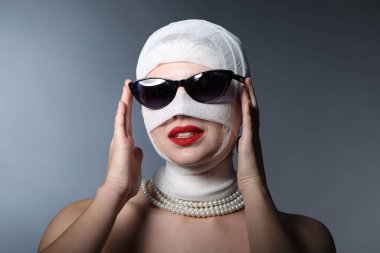 Gri arkaplandaki estetik ameliyattan sonra yüzü tıbbi bandaja sarılı bir kadın.