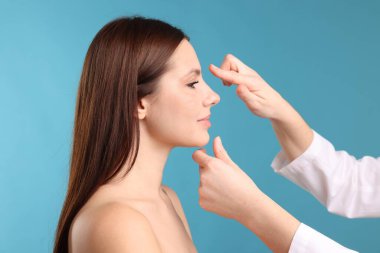 Açık mavi arka planda estetik ameliyattan önce hastanın yüzünü kontrol eden doktor, yakın plan.