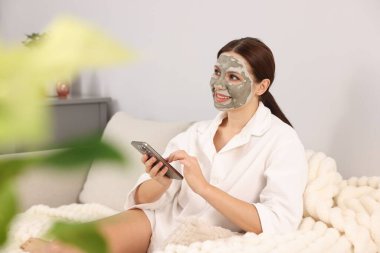 Yüzünde kilden maske olan bir kadın evde akıllı telefon kullanıyor.