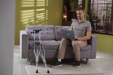 Evdeki koltuk değneklerinin yanındaki koltukta dizüstü bilgisayar kullanan bir adam.