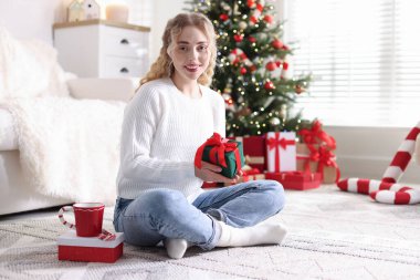 Evde bulanık ışıklara karşı elinde Noel hediyeleriyle gülümseyen bir kadın.