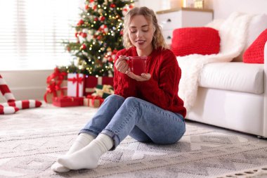 Süveter giyen, elinde bir bardak sıcak içecekle gülümseyen kadın Noel için dekore edilmiş odadaki bulanık ışıklara karşı.