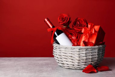 Şarap, şeker, hediye kutusu ve güllerle dolu sepet kırmızı arka planda gri masada, mesaj için yer var.
