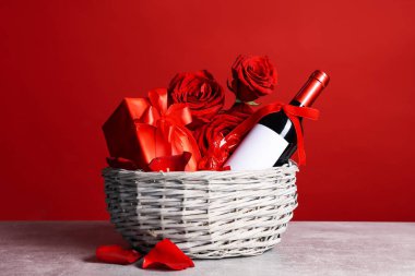Kırmızı arka planda şarap, şeker, hediye kutusu ve güllerle dolu bir sepet.