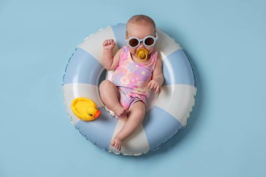 Güneş gözlüklü, oyuncak ördekli, şişme yüzüklü, açık mavi arka planda şirin bir bebek.
