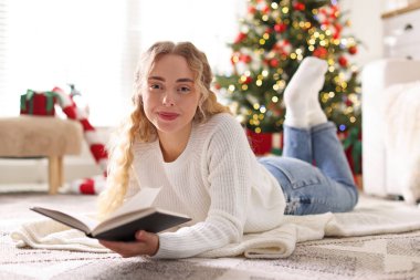Süveter giymiş güzel bir kadın, yerde kitap var. Odadaki bulanık ışıklara karşı, Noel için dekore edilmiş.