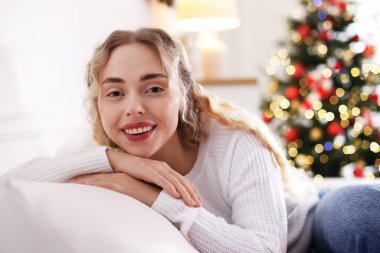 Evdeki bulanık ışıklara karşı kanepede süveter giymiş gülümseyen kadın portresi. Mutlu Noeller ve mutlu yıllar.