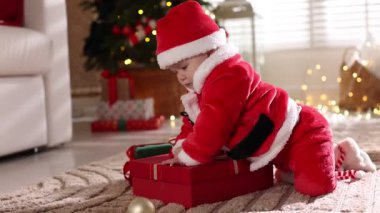 Noel Baba kostümlü şirin bir bebek. Evde yerde Noel hediyeleri var.