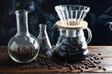 Kahve, fasulye ve labaratuar bardaklı cam kahve makinesi ahşap masada siyah arka planda, yakın planda.