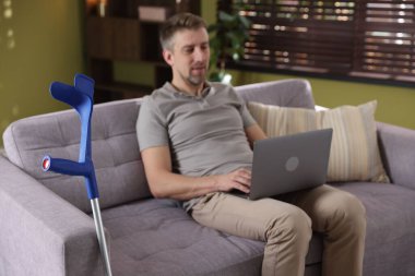 Evdeki koltukta dizüstü bilgisayar kullanan adam, dirsek değneğine odaklan.