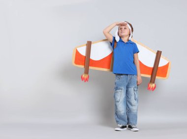 Kartondan uçak kanatları olan küçük çocuk açık gri arka planda bir şeye bakıyor. Metin için boşluk