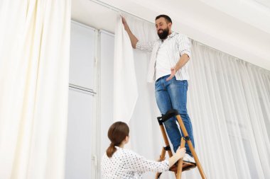 Evde pencere perdelerini asan merdivenli çift, düşük açı manzaralı.