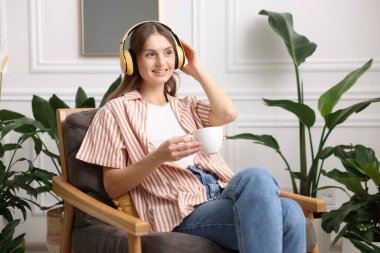 Elinde bir fincan aromatik kahve olan kadın evdeki koltukta müzik dinliyor.