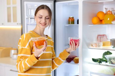 Mutfaktaki açık buzdolabının yanında yiyecek ürünleri olan kadın.