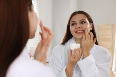 Bornozlu kadın evin içinde aynanın yanında yüzüne krem sürüyor.