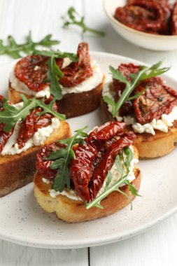 Krem peynirli lezzetli bruschettalar, güneşte kurutulmuş domatesler ve beyaz masada roka, yakın plan.