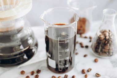 Lezzetli kahve, fasulye, toz, laboratuvar bardağı ve kimya kahve makinesi beyaz mermer masada, yakın plan.