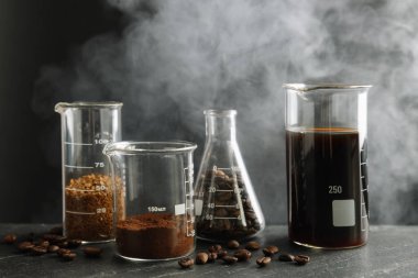 Lezzetli kahve, fasulye, toz ve dumanlı laboratuvar bardakları siyah masada, yakın plan.