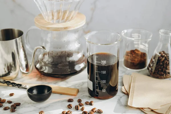 Lezzetli kahve, fasulye, labaratuvar bardağı, su ısıtıcısı, kağıt filtreler ve beyaz mermer masada kimya kahve makinesi, yakın plan.