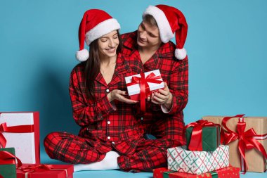 Noel Baba şapkalı ve pijamalı mutlu çift açık mavi arka planda Noel hediyeleri