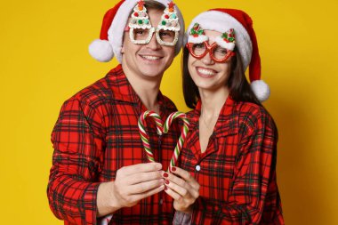 Noel Baba şapkalı ve pijamalı mutlu çift turuncu arka planda şeker kamışlarıyla kalp yapıyorlar. Noel atmosferi