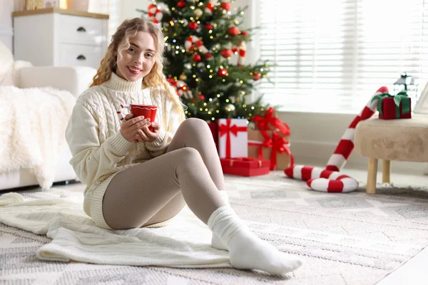 Süveter giymiş, elinde bir fincan sıcak içecekle gülümseyen bir kadın Noel için dekore edilmiş odanın bulanık ışıklarına karşı. Metin için boşluk