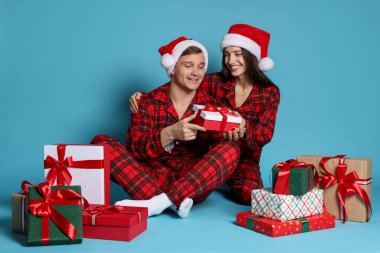 Noel Baba şapkalı ve pijamalı mutlu çift açık mavi arka planda Noel hediyeleri