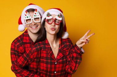 Noel Baba şapkalı mutlu çift, turuncu arka planda komik gözlükler ve pijamalar. Noel atmosferi