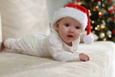 Noel Baba şapkalı şirin bir bebek Noel ışıklarına karşı kanepede, bokeh etkisi