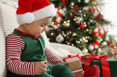 Little boy in Santa hat with gift boxes on sofa in room decorated for Christmas