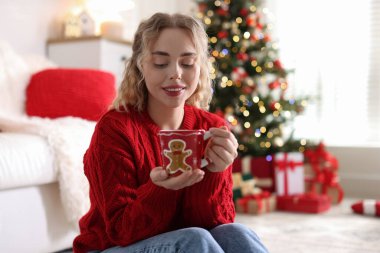 Süveter giymiş, elinde bir bardak sıcak içecekle gülümseyen bir kadın. Odadaki bulanık ışıklara karşı, Noel süslemesi yapılmış. Metin için boşluk