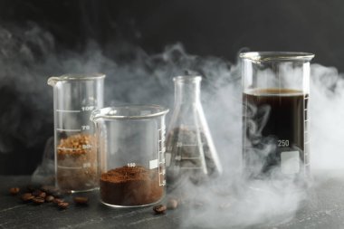 Lezzetli kahve, fasulye, toz ve dumanlı laboratuvar bardakları siyah masada, yakın plan.