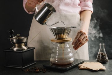 Kadın, siyah masadaki filtreli kimya kahve makinesine sıcak su döküyor. Siyah arka planda dumanlı, yakın plan.