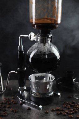 Vakumlu kahve makinesinde aromatik kahve demlemek, laboratuvar bardağı ve fasulye gri masada koyu arkaplanda dumanla kaplanmak, yakın plan