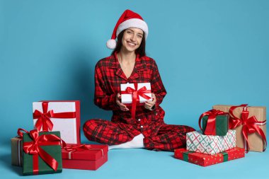 Noel Baba şapkalı ve pijamalı mutlu kadın. Açık mavi arka planda Noel hediyeleri.