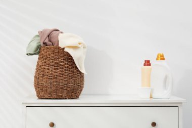 Çamaşır sepetinin içinde havlular ve deterjan şişeleri beyaz duvarın yanındaki dolapta.